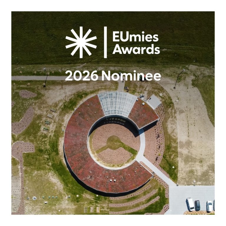 Zdjęcie z lotu ptaka okrągłego budynku z czerwonym, zielonym i szklanym dachem, otoczonego ścieżkami i zielenią. Na górze grafiki logo EUmies Awards i napis '2026 Nominee'