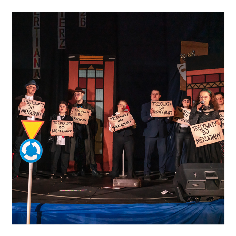Grupa osób stoi na scenie podczas przedstawienia teatralnego i trzyma kartki z napisem Trędowaty, bo niekochany, na tle scenografii miejskich kamienic